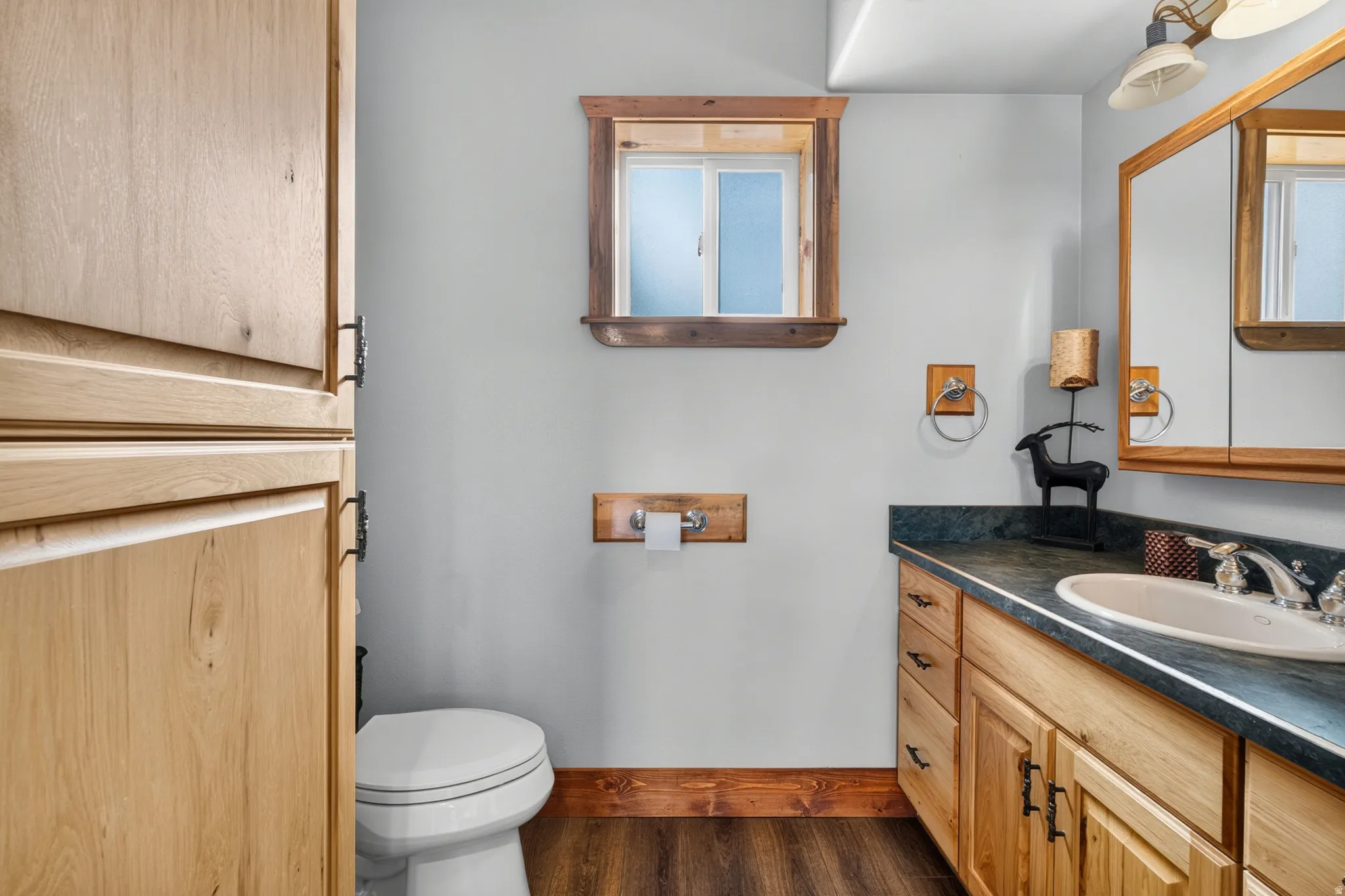 Bathroom featuring a wood-finish vanity with a built-in sink and dark countertop, wood-finish flooring, a toilet, a privacy window with a wood frame, and light-toned walls