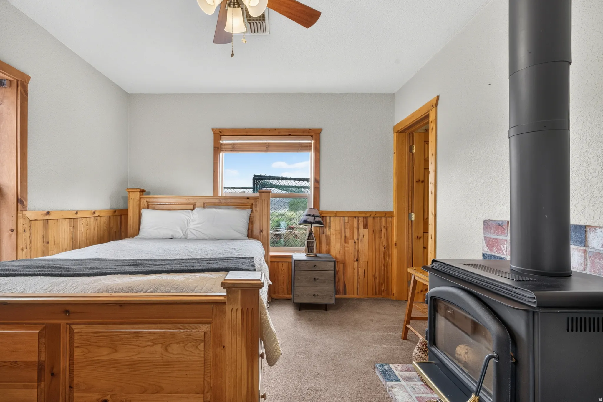 Room featuring natural wood wainscoting and trim, a corner wood-burning stove with a brick-finish hearth, a ceiling fan with integrated lighting, and carpet flooring