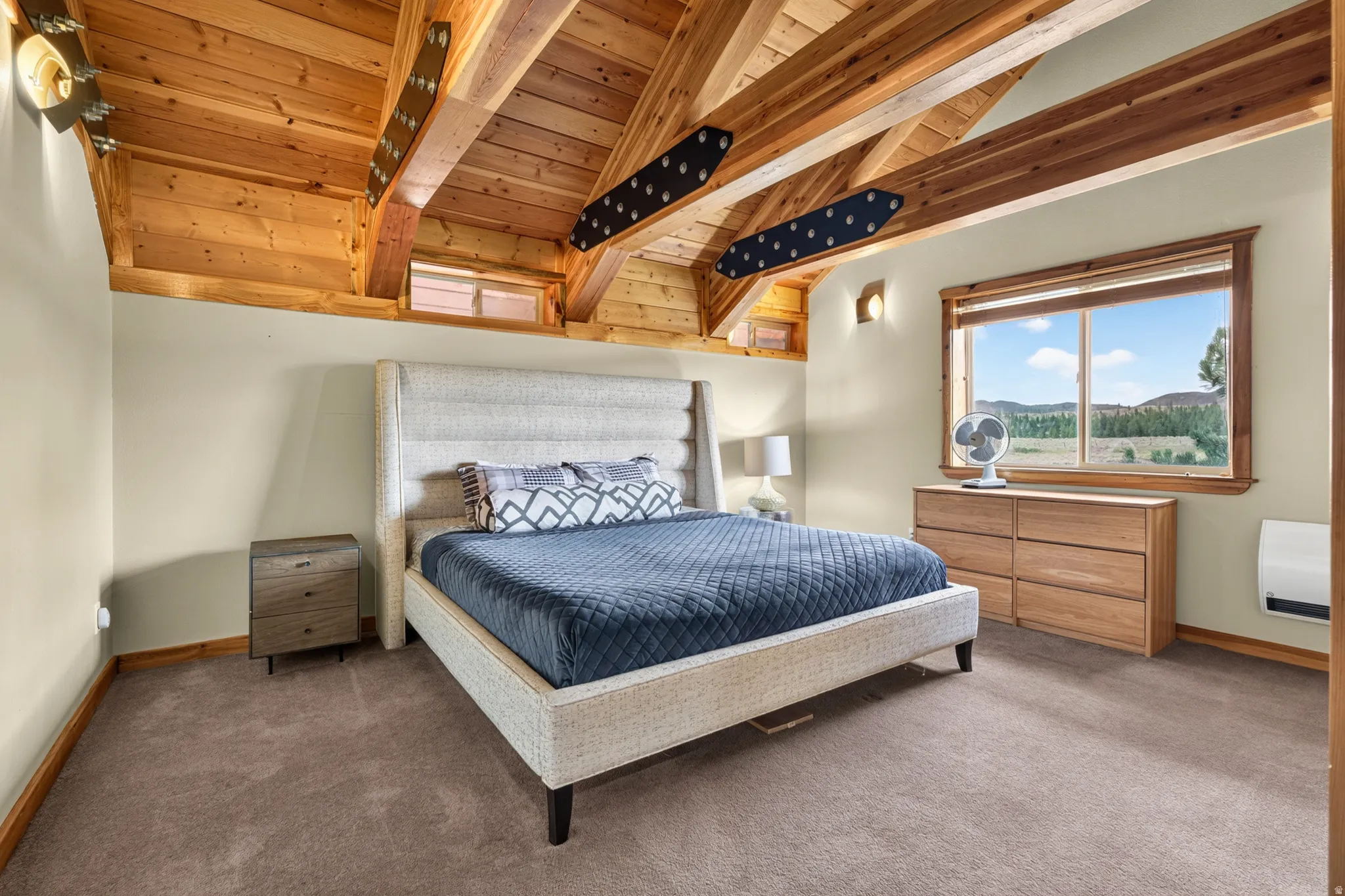 Vaulted wood-paneled ceiling with exposed beams, wood-trimmed window, wall-mounted lighting fixtures, and light-toned carpet flooring