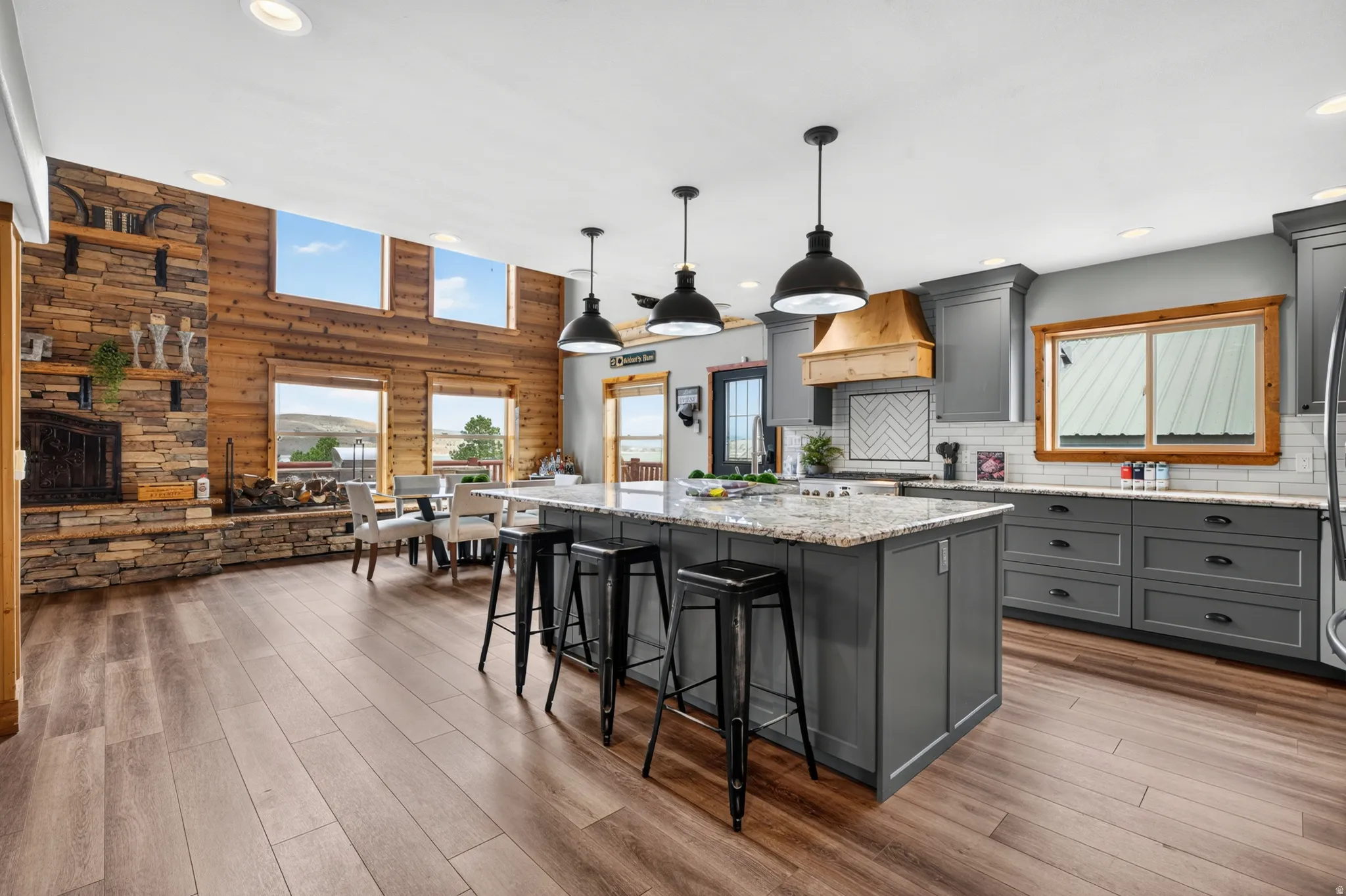 Open-concept living space featuring wood-finish flooring, a stone fireplace with a wood mantle, and a kitchen island with a stone-finish countertop