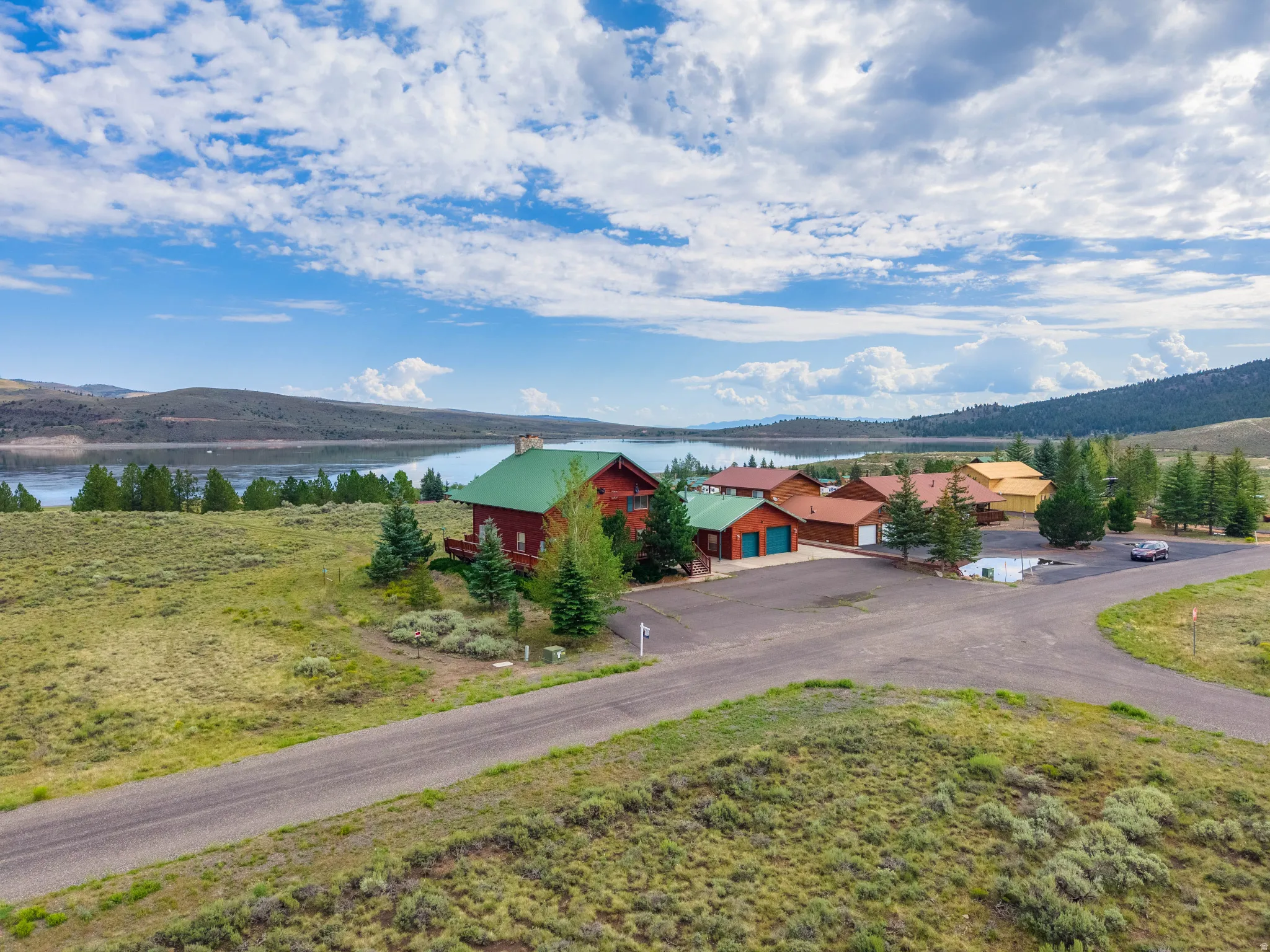 Expansive property featuring a red-sided structure with a green roof, multiple outbuildings, mature evergreen trees, and a paved driveway, all situated against a scenic lake and mountainous backdrop