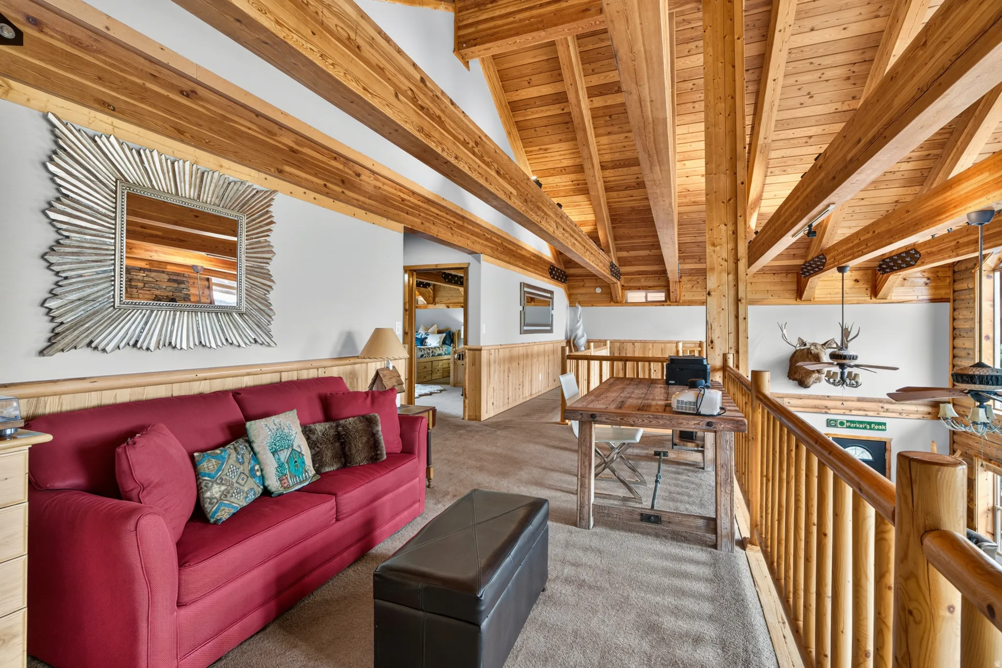 Loft space featuring exposed natural wood beams and ceiling, log-style railings, wall-mounted light fixtures, and a neutral carpeted floor