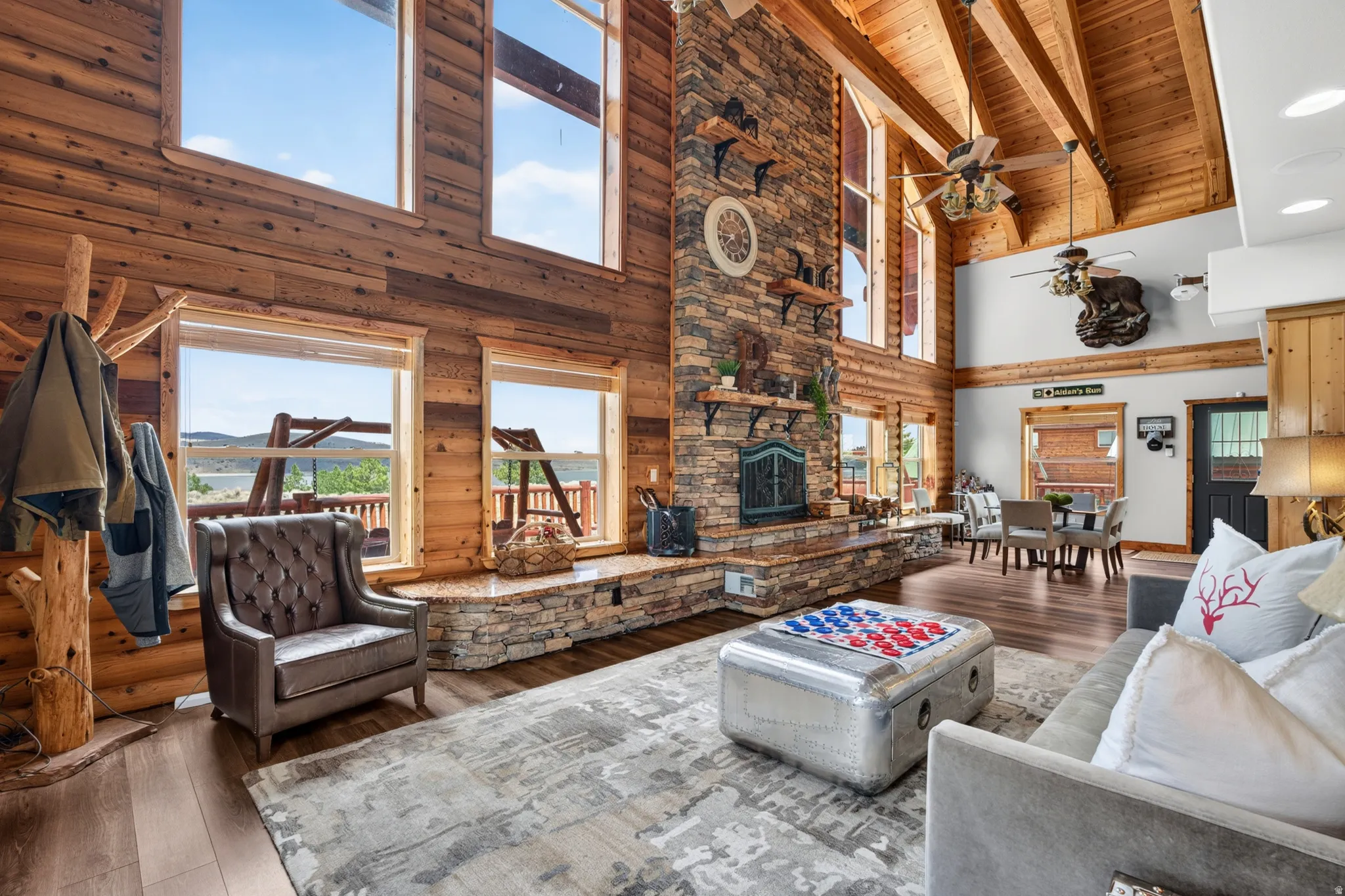 Spacious living area featuring wood-finish flooring, a stacked stone fireplace with a stone hearth, exposed wood beams, and multiple large windows