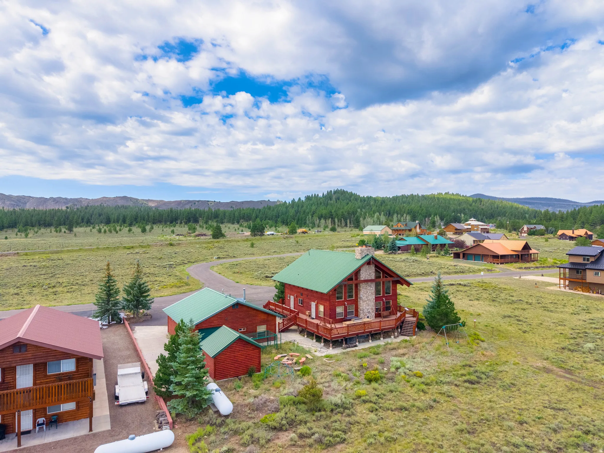 Expansive property featuring a red log cabin with a green roof, a stone-faced chimney, and a surrounding wooden deck