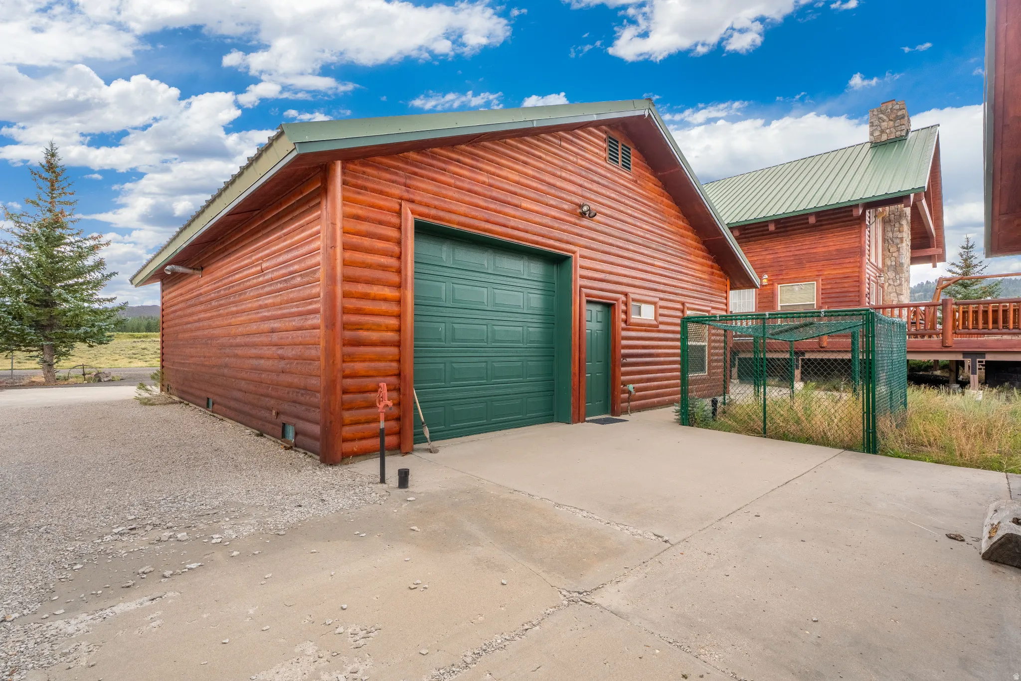 Detached log-style garage featuring a green garage door and personnel door, concrete driveway, and gravel parking area