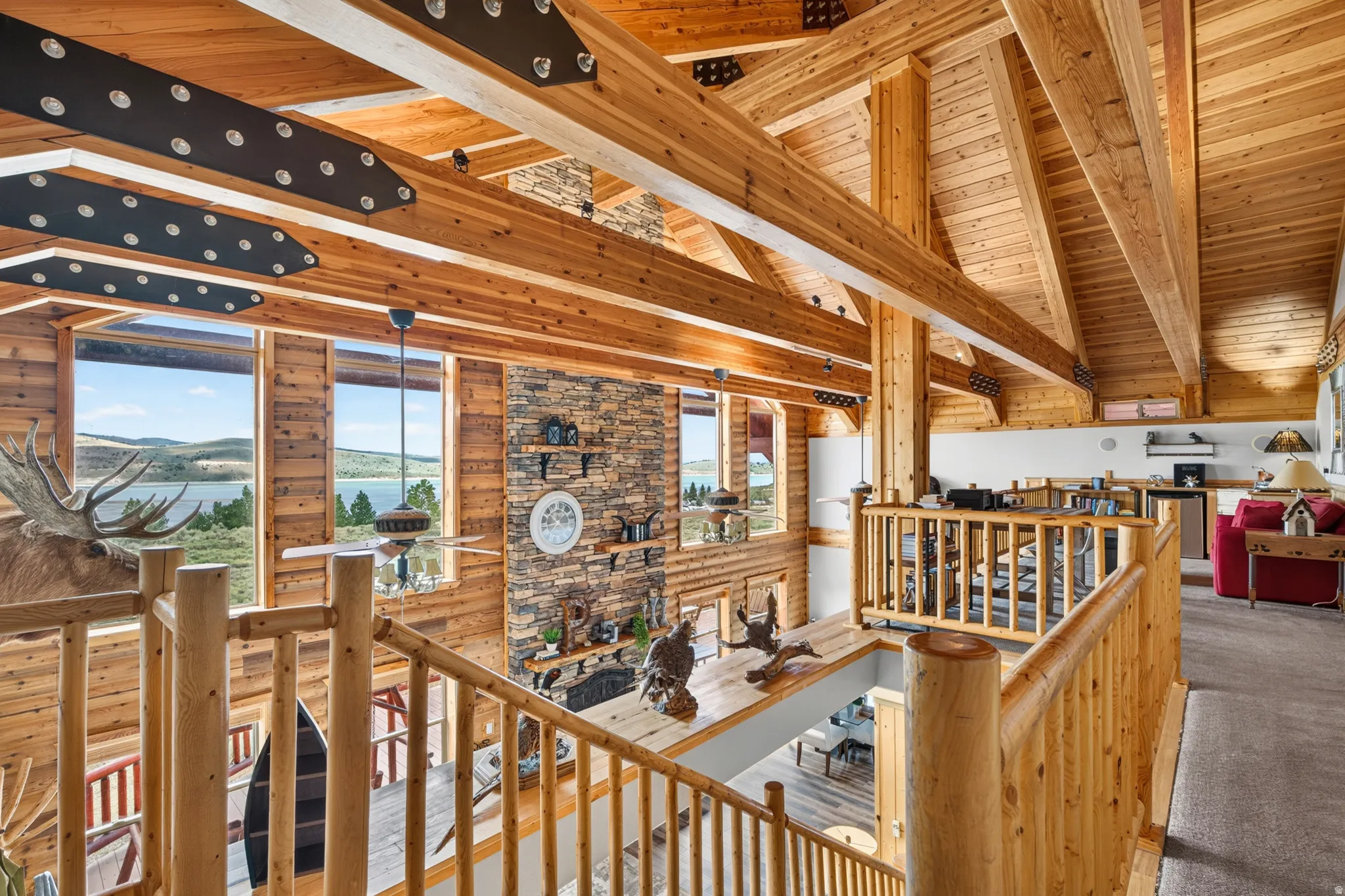 Expansive interior featuring exposed timber beams, vaulted wood ceilings, a stone accent wall, large windows, and an upper-level overlook