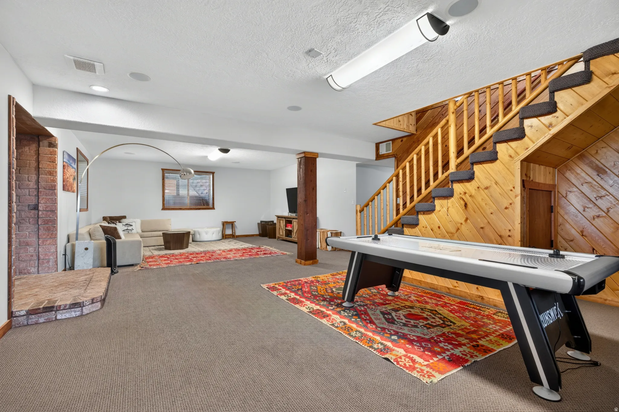 Expansive recreation room featuring a natural wood staircase with carpeted treads, wood paneling, recessed lighting, and a brick fireplace
