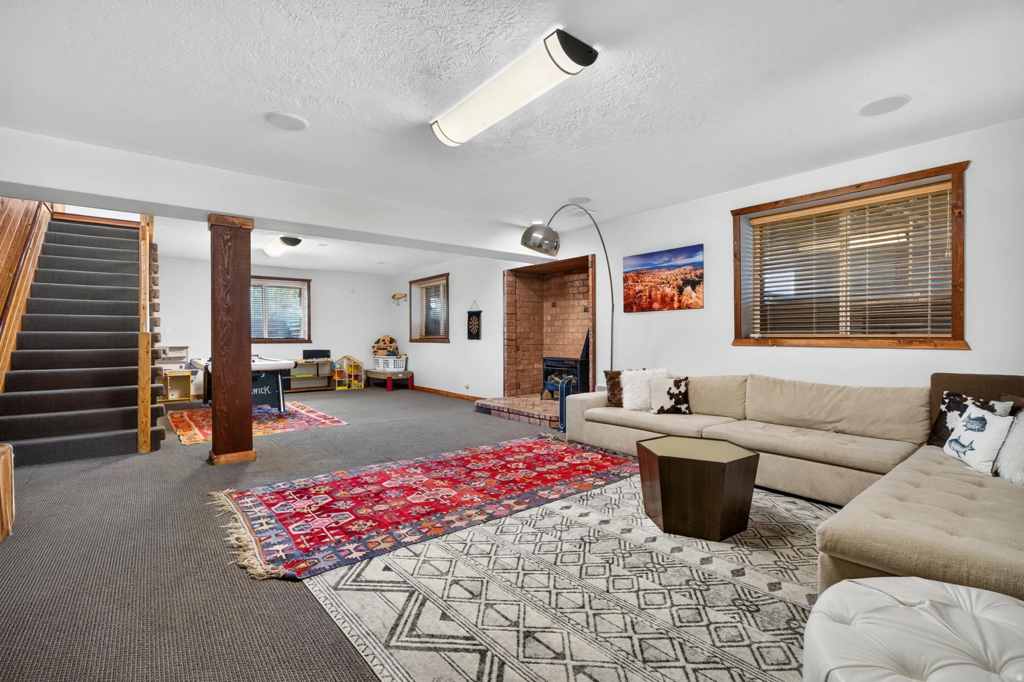 Spacious room featuring gray carpet flooring, wood-framed window with blinds, and a brick fireplace
