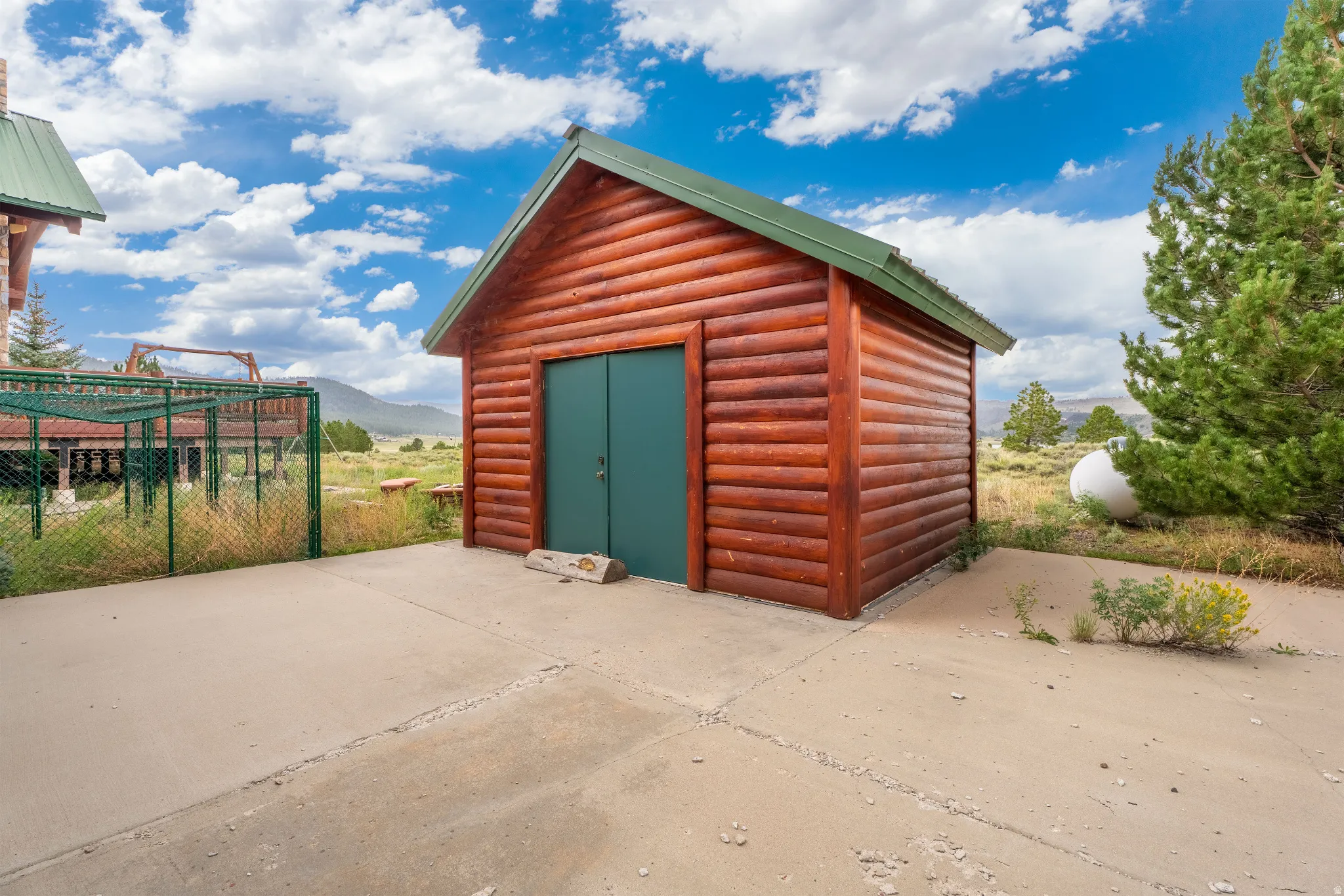 Log cabin-style outbuilding with a green metal roof and matching double doors, positioned on a concrete pad