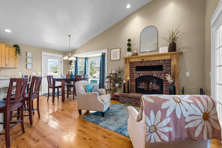 Open-concept living space featuring hardwood flooring, a wood-burning fireplace with brick surround and arched mantel, recessed ceiling lighting, and large windows with natural light
