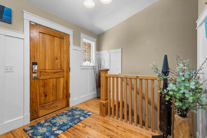 Welcoming foyer showcasing hardwood flooring, a solid wood interior door with hardware detail, wooden stair railing with vertical spindles, recessed ceiling lighting, neutral wall finishes, and decorative potted greenery