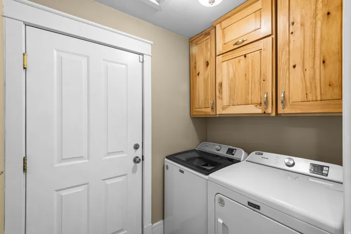 Dedicated laundry room featuring white front-load washer and dryer with digital controls, ample wood-finish cabinetry for storage, neutral wall finishes, and direct entry from the main living areas