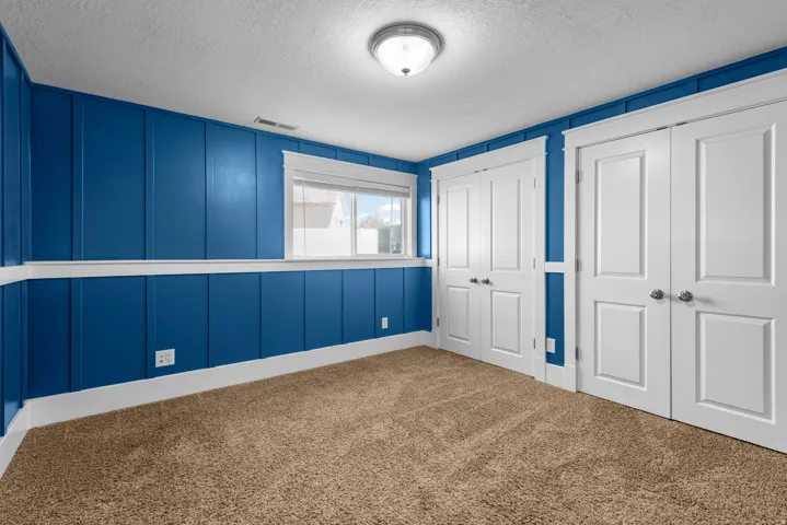 Spacious bedroom featuring bold blue panel wainscoting with white trim detail, neutral carpet flooring, dual white six-panel closet doors, and large window providing natural light