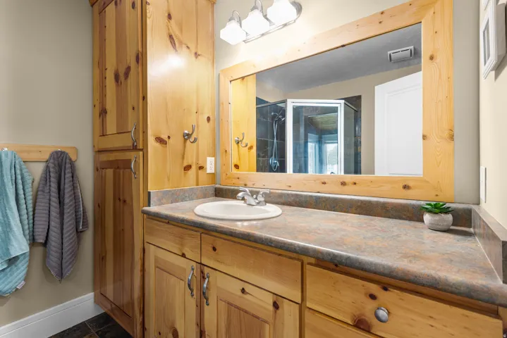 Spa-inspired master bathroom featuring knotty pine cabinetry with ample storage, dual-sink vanity with stone countertop, large framed mirror with integrated lighting, and brushed nickel hardware