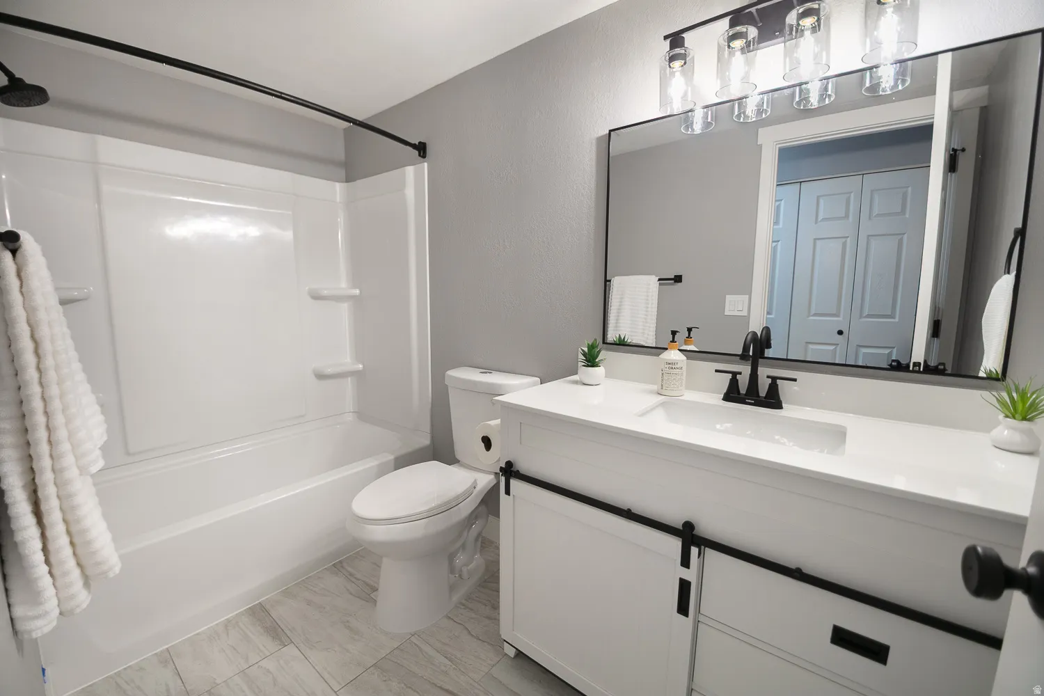 Bathroom featuring a white bathtub with integrated shower, white vanity with barn door style cabinetry, integrated sink, and dark-finish fixtures, a large rectangular mirror, and light-toned tile flooring