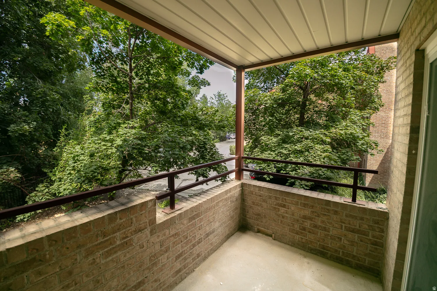Covered exterior balcony featuring a neutral-toned brick half-wall, dark metal railing, and a slatted ceiling
