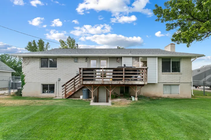 Expansive backyard featuring a manicured lawn, a two-story residence with a light-colored brick and siding exterior, a large wood deck with stairs, a ground-level basement entrance, and a gray shingle roof