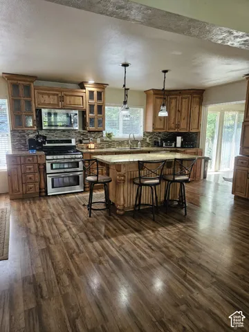 Spacious kitchen featuring wood-finish flooring, custom wood cabinetry, stainless steel appliances, a multi-toned tile backsplash, and a large center island with pendant lighting