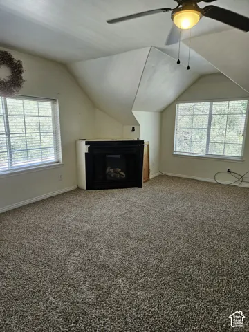 Carpeted interior space featuring a built-in fireplace, two windows with blinds, a ceiling fan with integrated lighting, and sloped ceiling details
