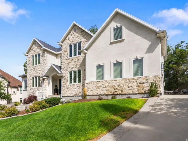 Two-story property featuring a stone and stucco exterior, gabled rooflines, a concrete driveway, and a landscaped front yard with mature trees