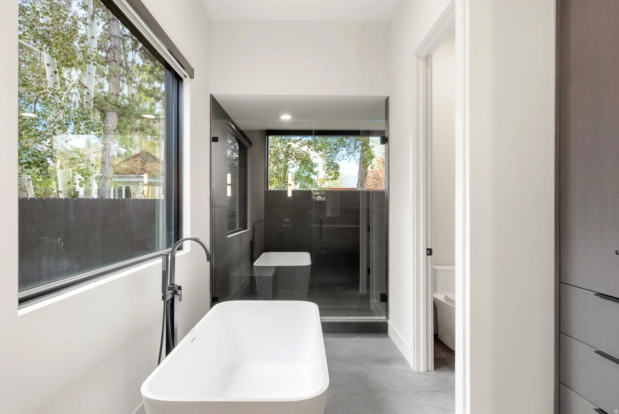 Contemporary bathroom featuring a freestanding soaking tub with a floor-mounted matte black faucet, expansive windows providing natural light, a glass-enclosed shower, and built-in cabinetry with modern hardware