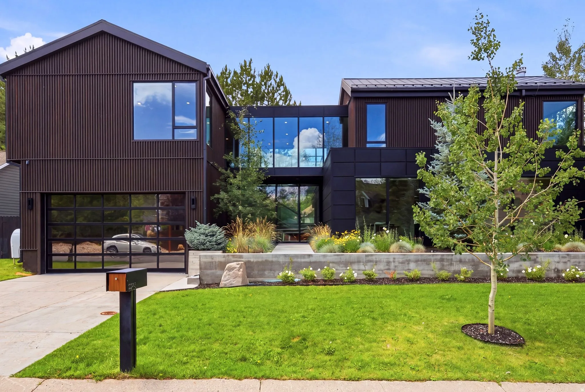 Modern architectural residence with dark vertical panel siding, extensive floor-to-ceiling windows, a full-view garage door, a concrete retaining wall with integrated landscaping, and a manicured front lawn