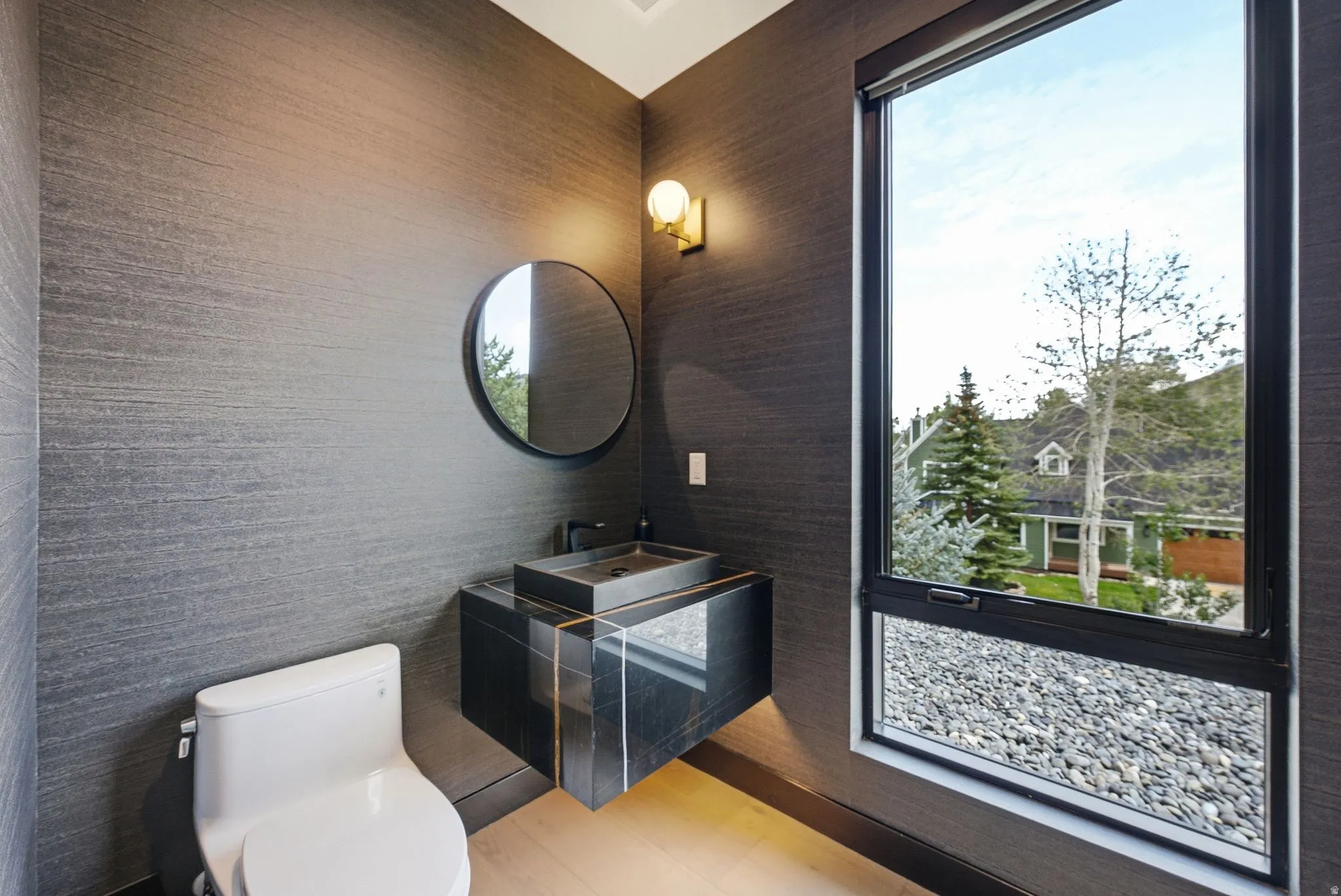Powder room with textured dark gray wall covering, a floating vanity featuring a dark stone countertop and vessel sink, a circular mirror, and a wall-mounted light fixture