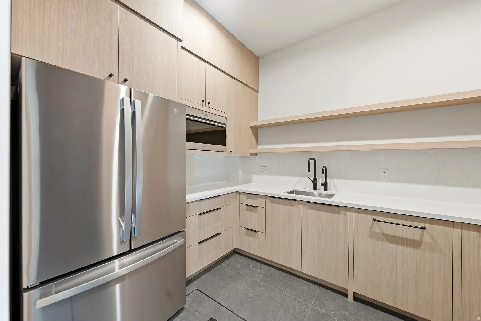 Modern kitchen space featuring a stainless steel French door refrigerator, integrated microwave, light wood-finish cabinetry with black hardware, white countertops, and a black matte faucet