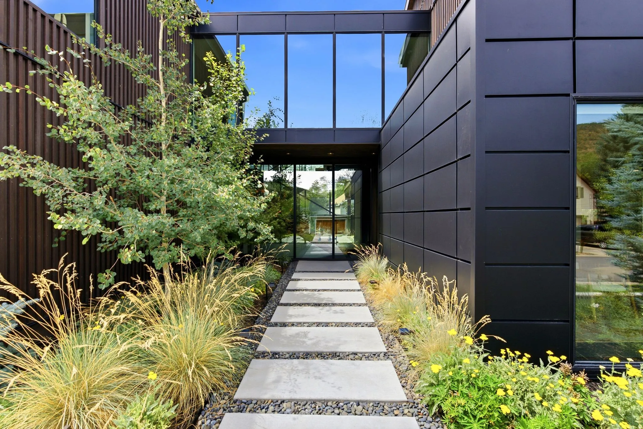 Contemporary architectural design featuring a glass-enclosed entry bridge, dark paneling, a concrete paver walkway, and integrated landscaping with ornamental grasses and mature plantings