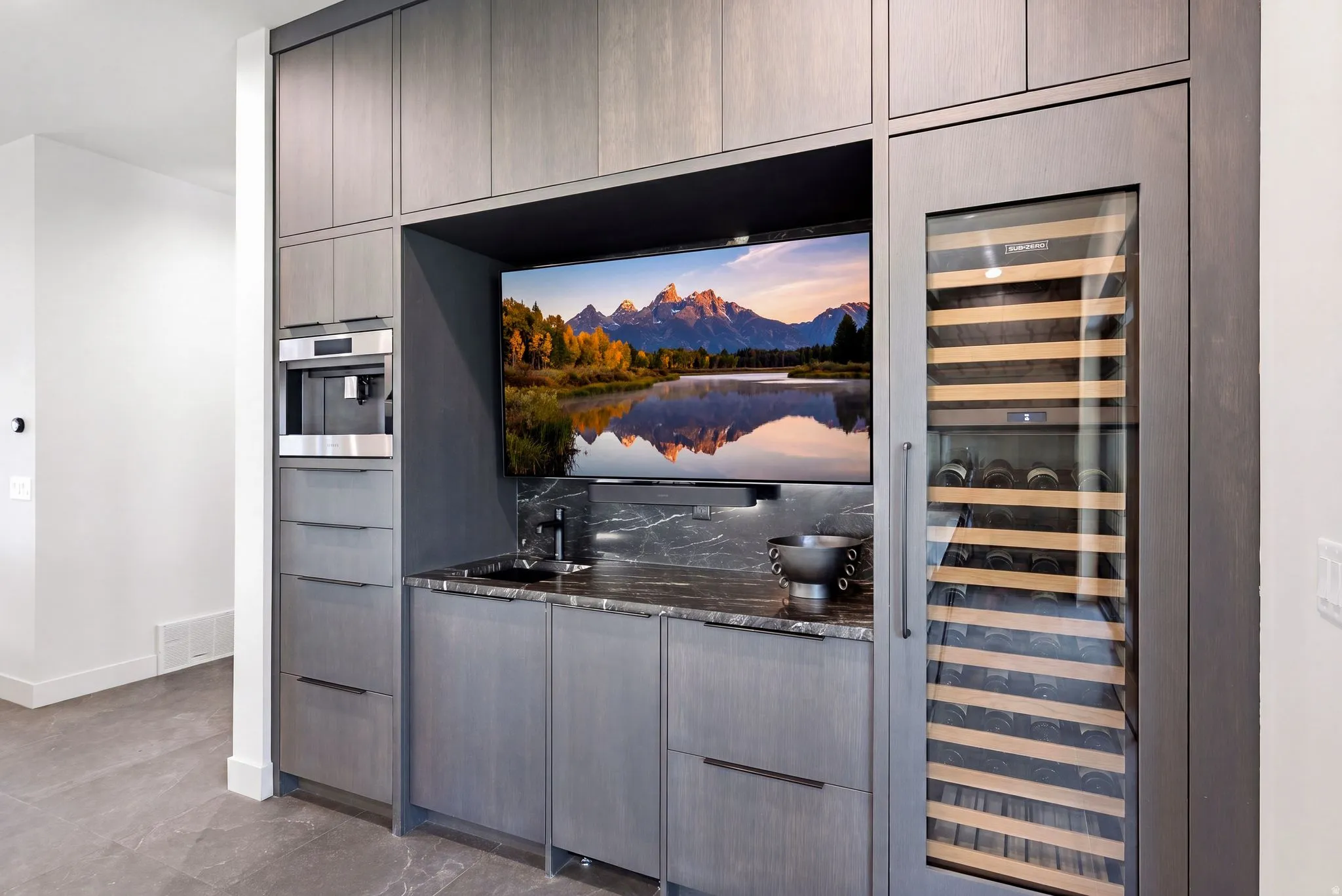 Integrated entertainment bar featuring wood-finish cabinetry, built-in television, undermount sink, stone countertop and backsplash, and an integrated beverage refrigerator