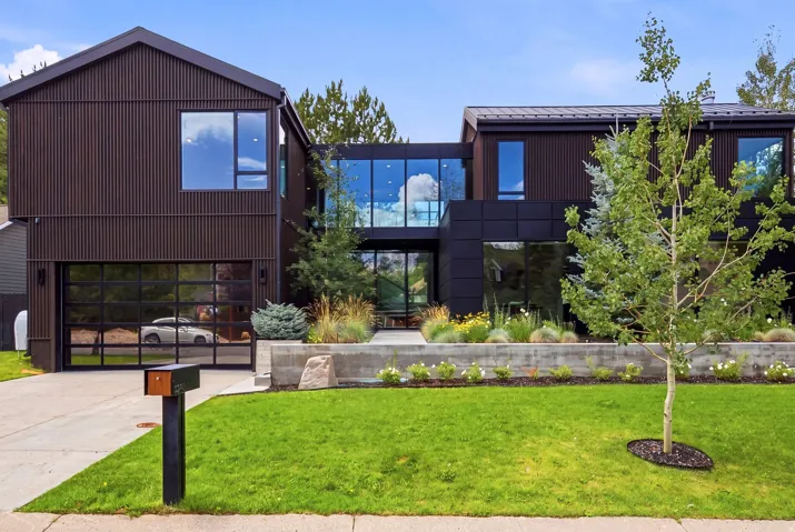 Modern architectural residence with dark vertical panel siding, extensive floor-to-ceiling windows, a full-view garage door, a concrete retaining wall with integrated landscaping, and a manicured front lawn