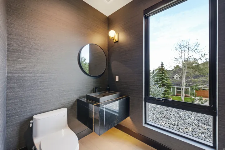 Powder room with textured dark gray wall covering, a floating vanity featuring a dark stone countertop and vessel sink, a circular mirror, and a wall-mounted light fixture