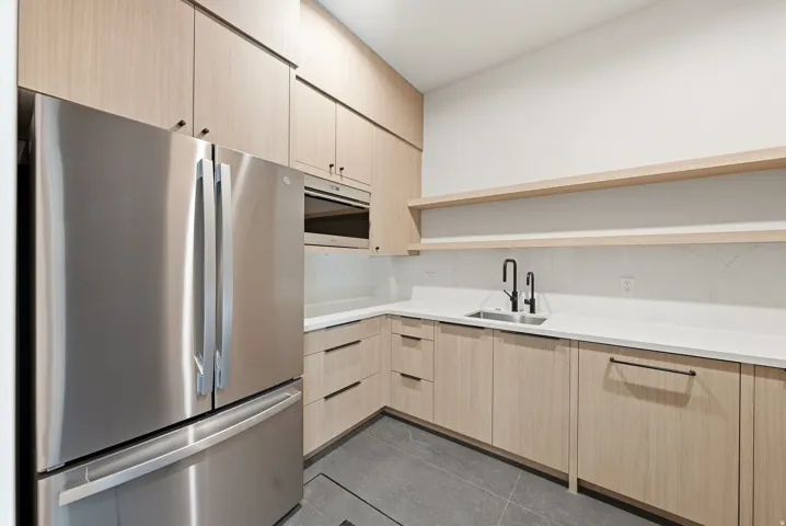 Modern kitchen space featuring a stainless steel French door refrigerator, integrated microwave, light wood-finish cabinetry with black hardware, white countertops, and a black matte faucet
