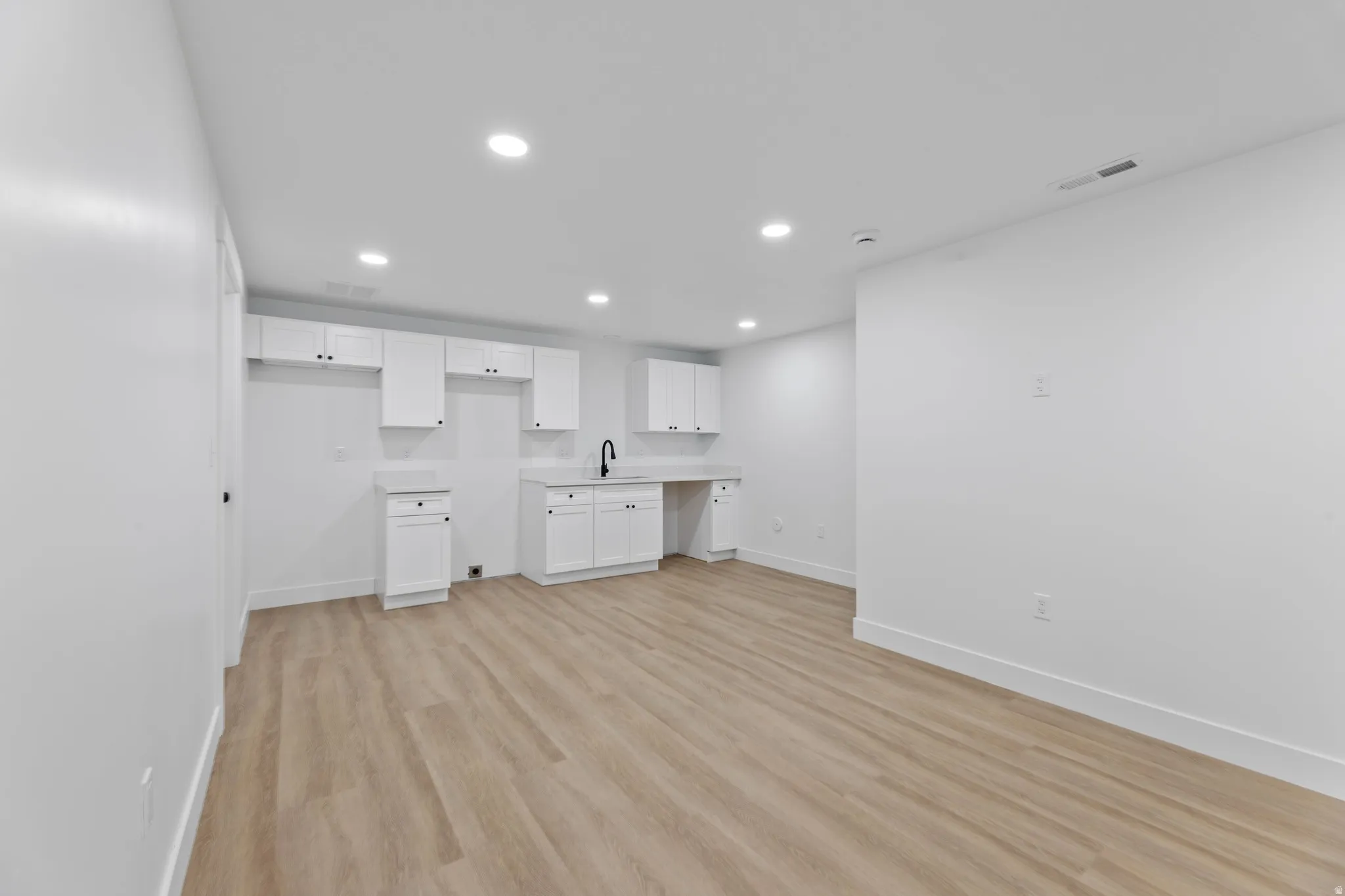 Interior space featuring wood-finish flooring, recessed lighting, white perimeter walls, white baseboards, and a built-in kitchenette area with white cabinetry, white countertops, and a black gooseneck faucet