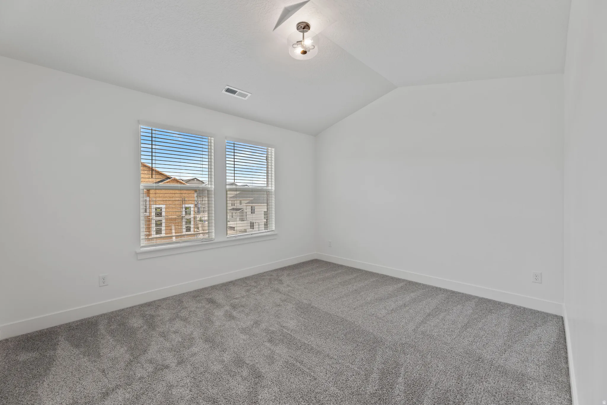 Bright room featuring a vaulted ceiling with a semi-flush mount light fixture, two large windows with blinds, plush gray carpeting, and white baseboards