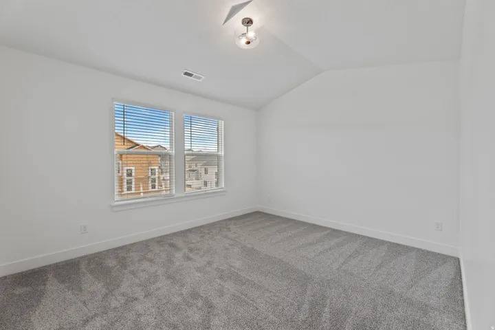 Bright room featuring a vaulted ceiling with a semi-flush mount light fixture, two large windows with blinds, plush gray carpeting, and white baseboards