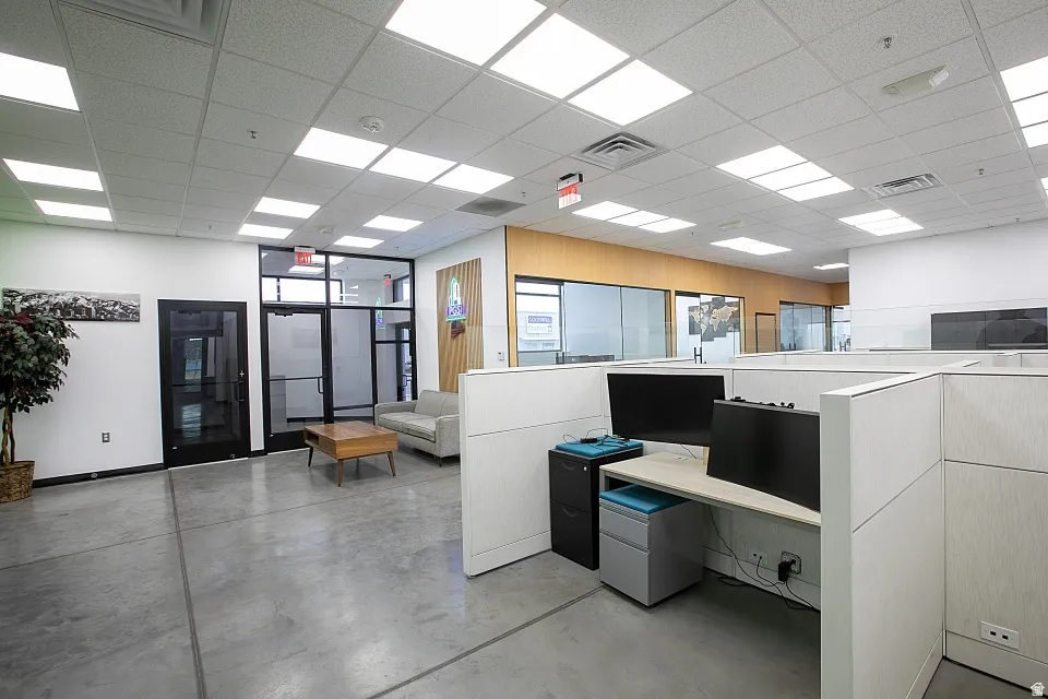 Open-plan office area featuring concrete flooring, a dropped ceiling with integrated rectangular lighting, and framed glass partitions