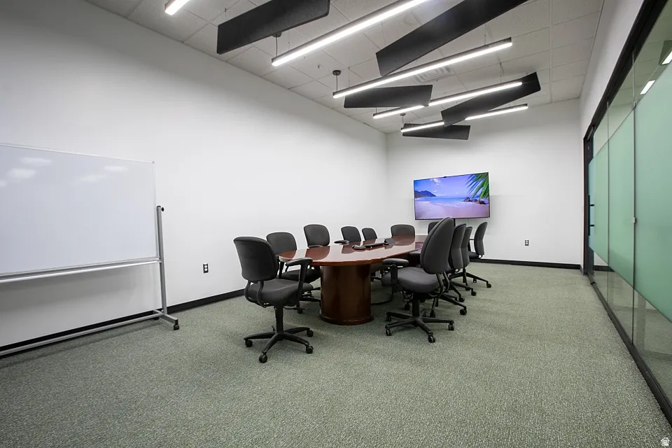 Conference room featuring a large mounted display, frosted glass wall, suspended acoustical panels, and linear pendant lighting
