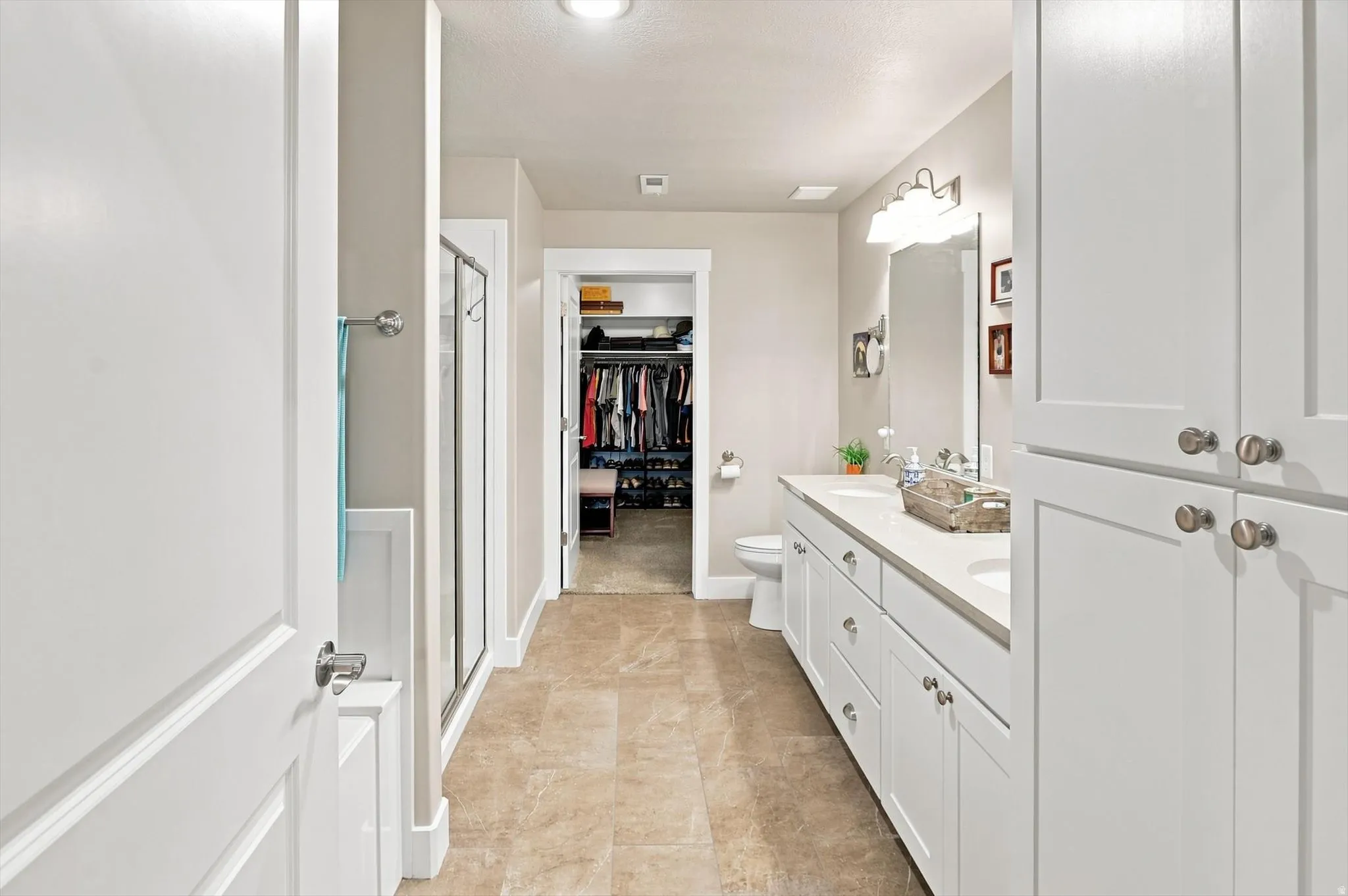Bathroom featuring dual vanity sinks with light countertops, extensive white cabinetry, a glass-enclosed shower, and neutral-toned tile flooring
