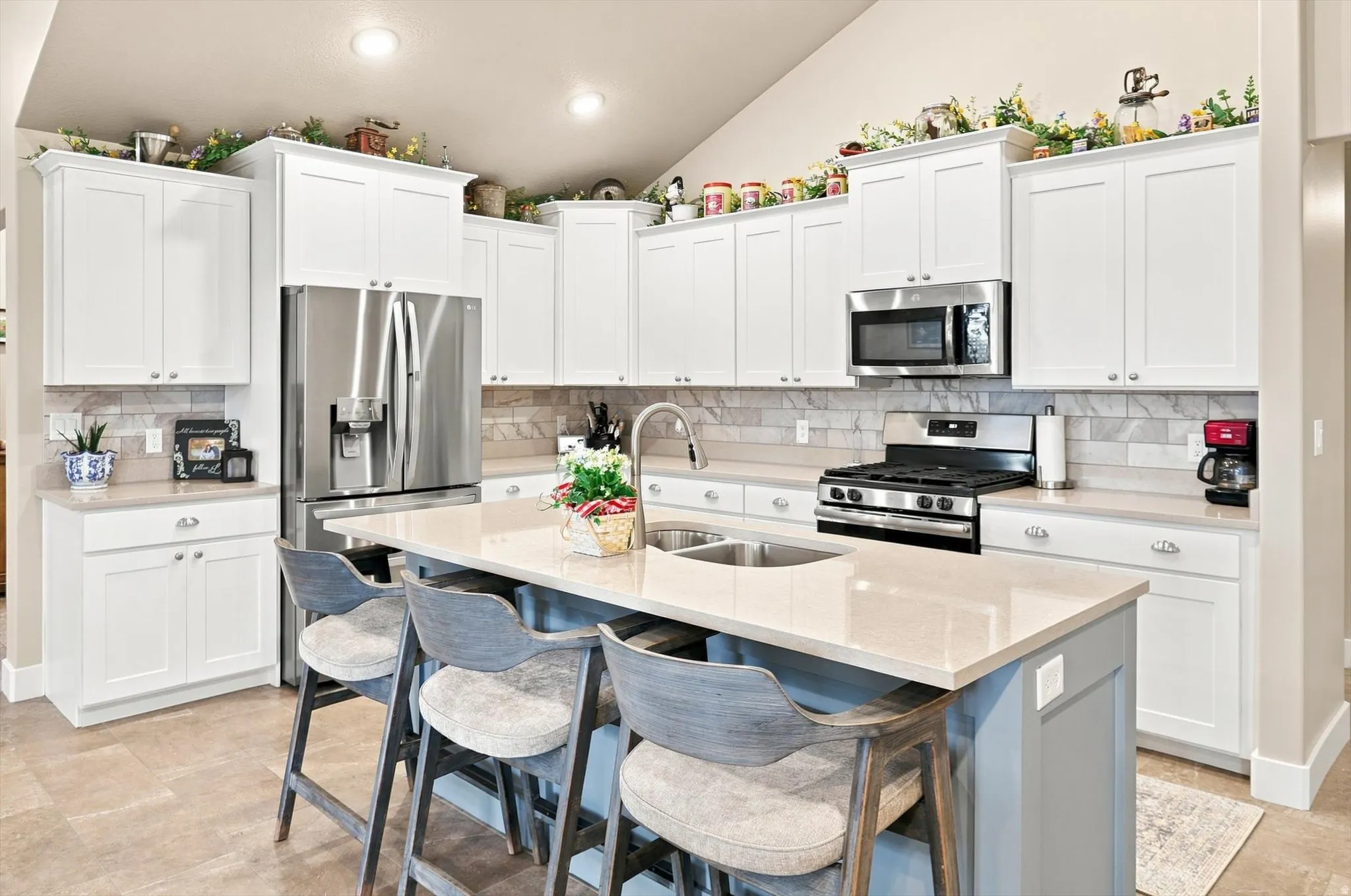 Open-concept kitchen featuring an island with a built-in sink, white shaker-style cabinetry, stainless steel appliances, a tile backsplash, and light-toned countertops