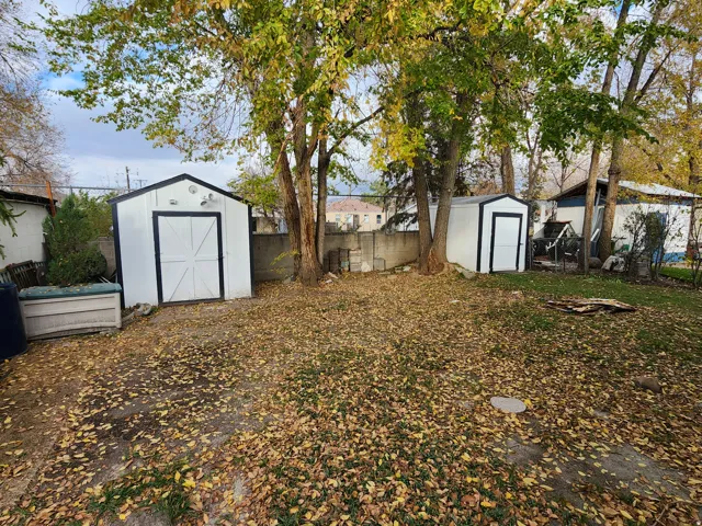 Expansive yard space featuring two white storage sheds with black trim, mature trees, and a concrete block retaining wall