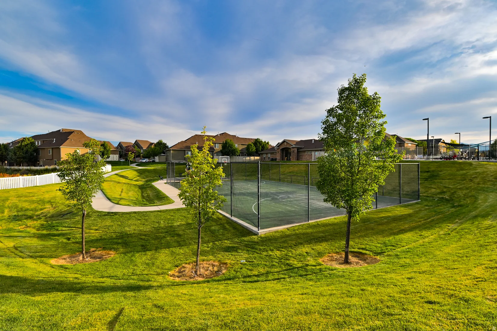 Community green space featuring a paved walking path, mature trees, and a fenced multi-sport court with basketball hoops and overhead lighting