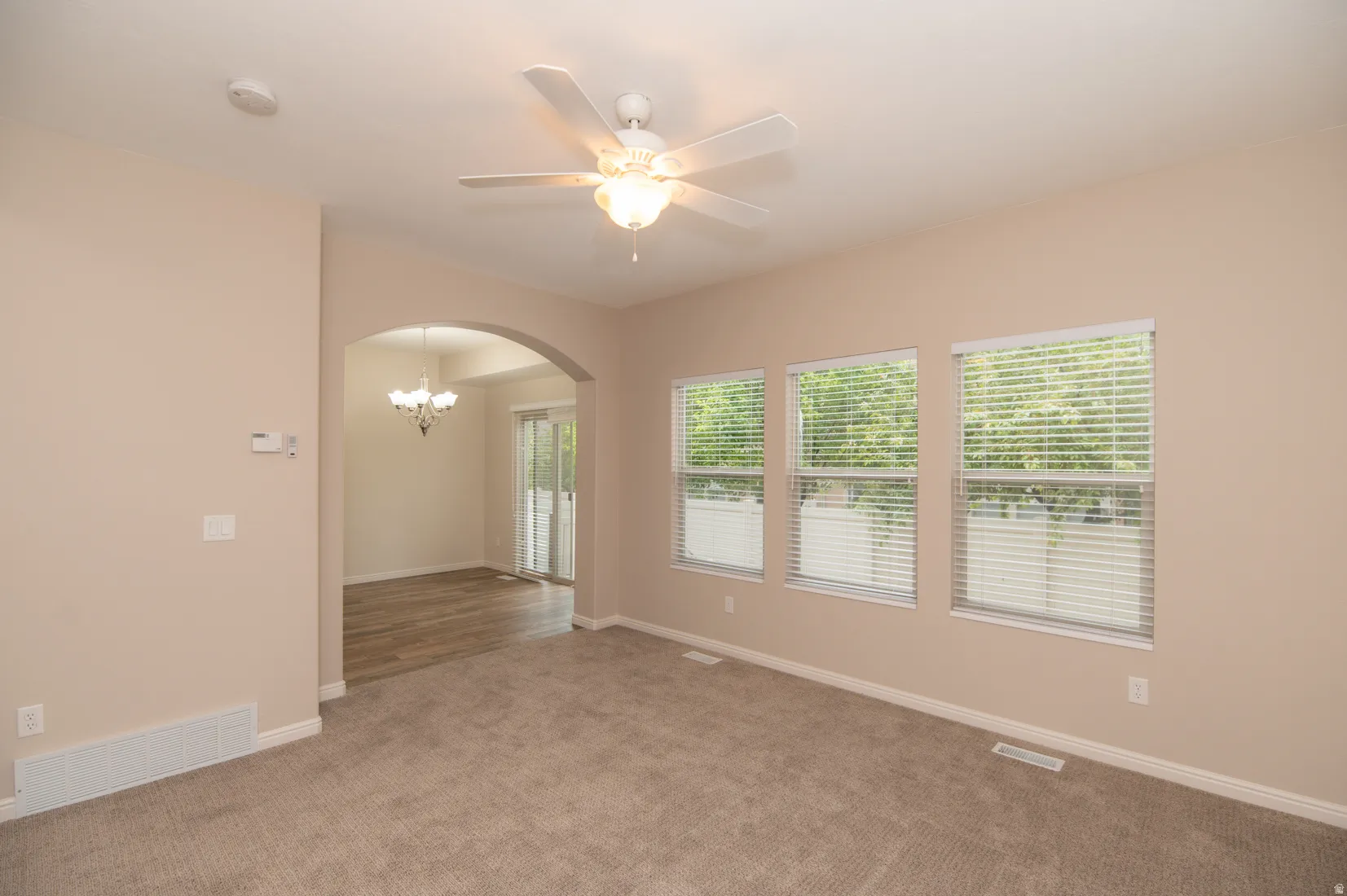 Carpeted living area featuring three windows with blinds, a ceiling fan with integrated lighting, and an arched doorway opening to a dining space