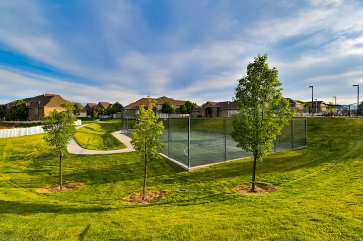 Community green space featuring a paved walking path, mature trees, and a fenced multi-sport court with basketball hoops and overhead lighting
