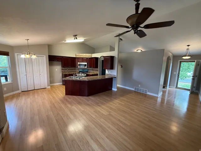 Open-concept living area featuring wood-finish flooring, a kitchen with dark wood cabinetry and stainless steel appliances, a central island with a sink, vaulted ceilings, and a ceiling fan