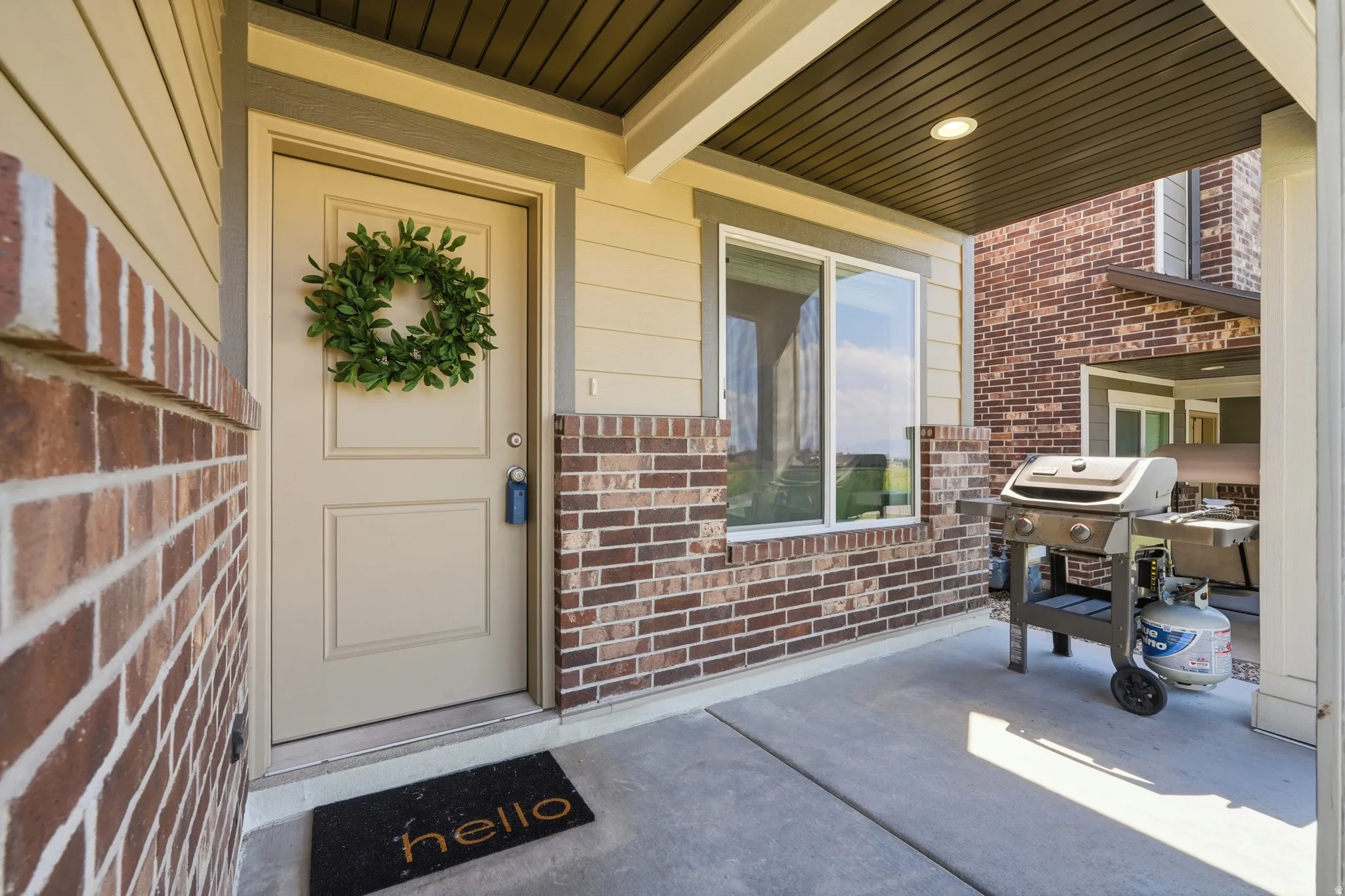 Covered front porch featuring a beige entry door, brick accents, horizontal siding, and a recessed light fixture