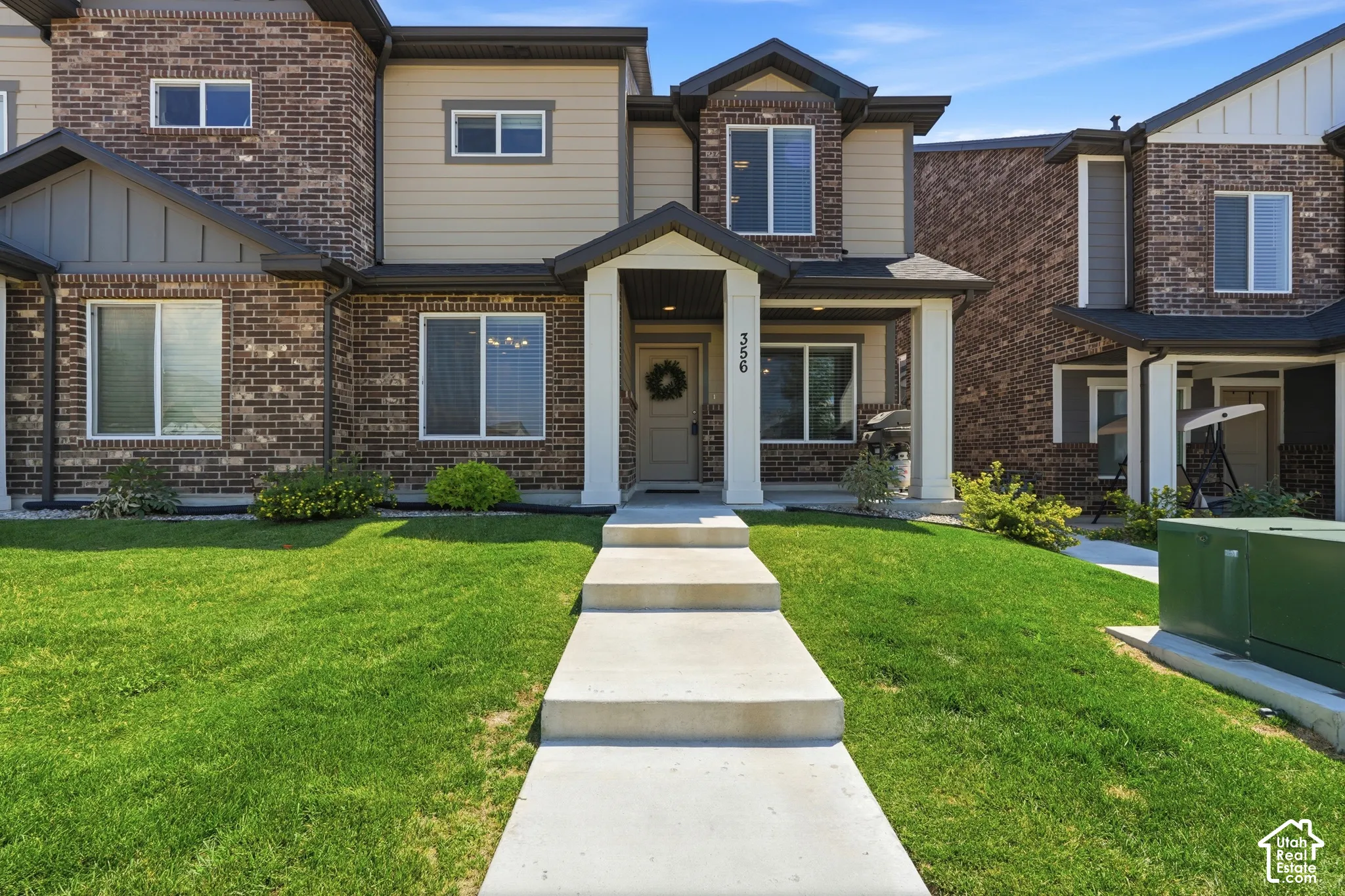 Brick and siding townhouse exterior featuring a covered porch with white columns, a concrete pathway, a manicured lawn, and multiple windows