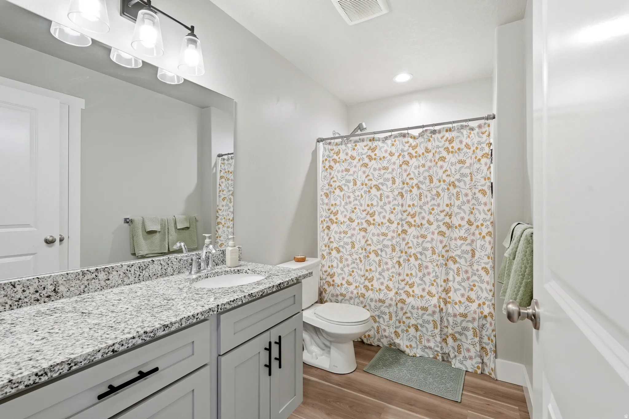 Bathroom featuring a light wood-finish floor, gray shaker-style vanity with matte black hardware, speckled granite countertop, undermount sink, and a shower/tub combination