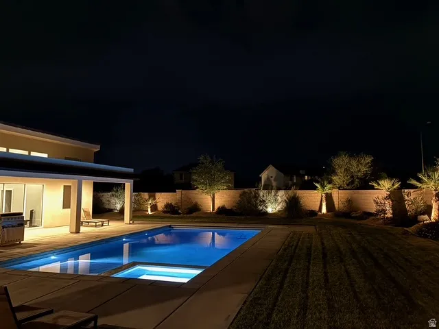 Backyard oasis featuring an illuminated swimming pool with an integrated spa, extensive concrete decking, a covered patio area, and perimeter landscaping with accent lighting