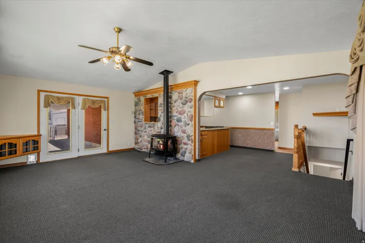 Spacious carpeted living area featuring a stone accent wall with a wood-burning stove, double glass French doors, and a ceiling fan